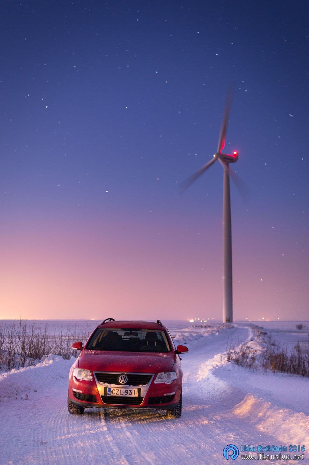 Punainen auto seisoo lumisella tiellä tuulivoimalan alla hämärässä