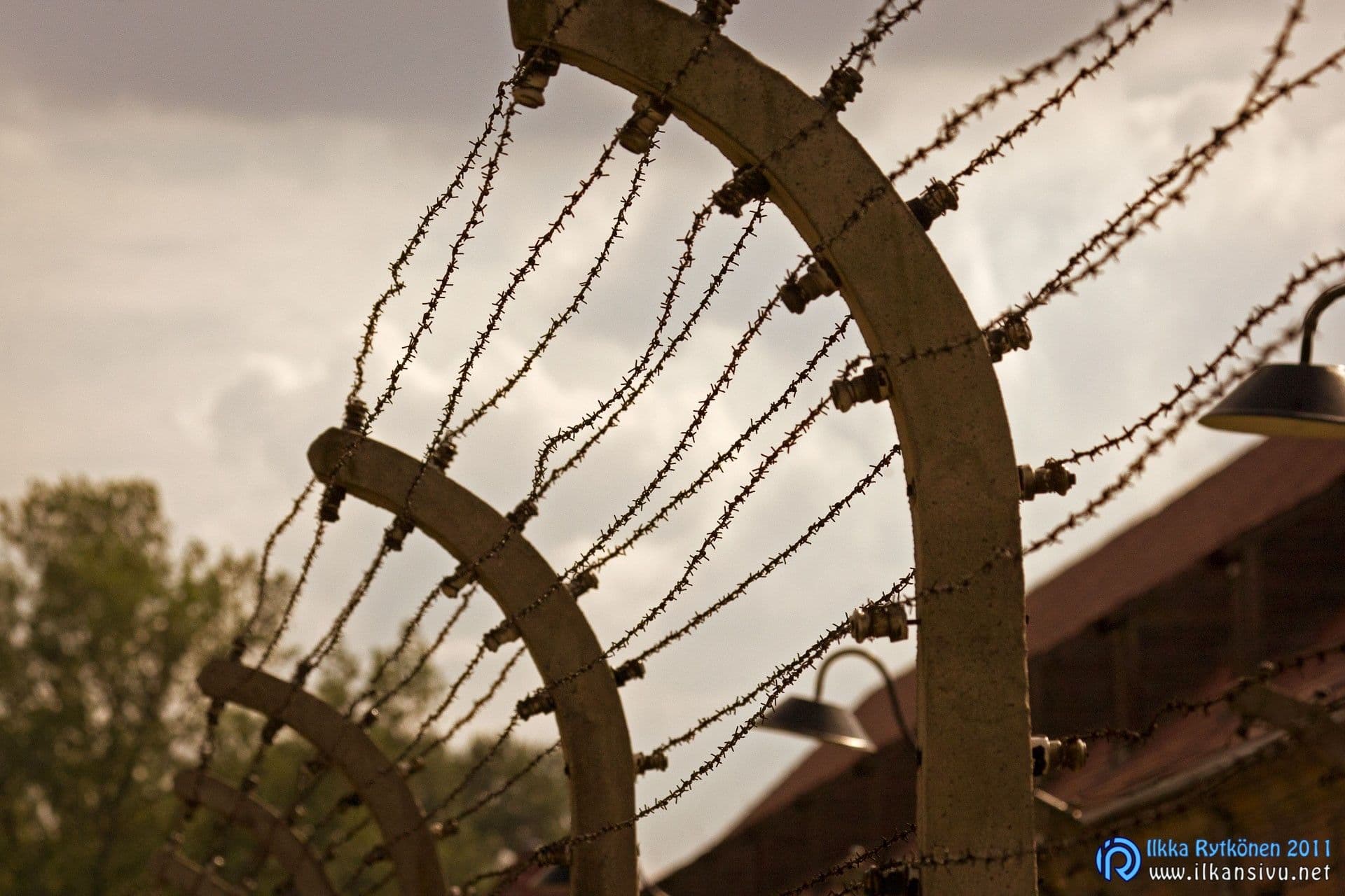 Auschwitz I:n sähköistetty piikkilanka-aita vastavalossa pilvistä taivasta vasten