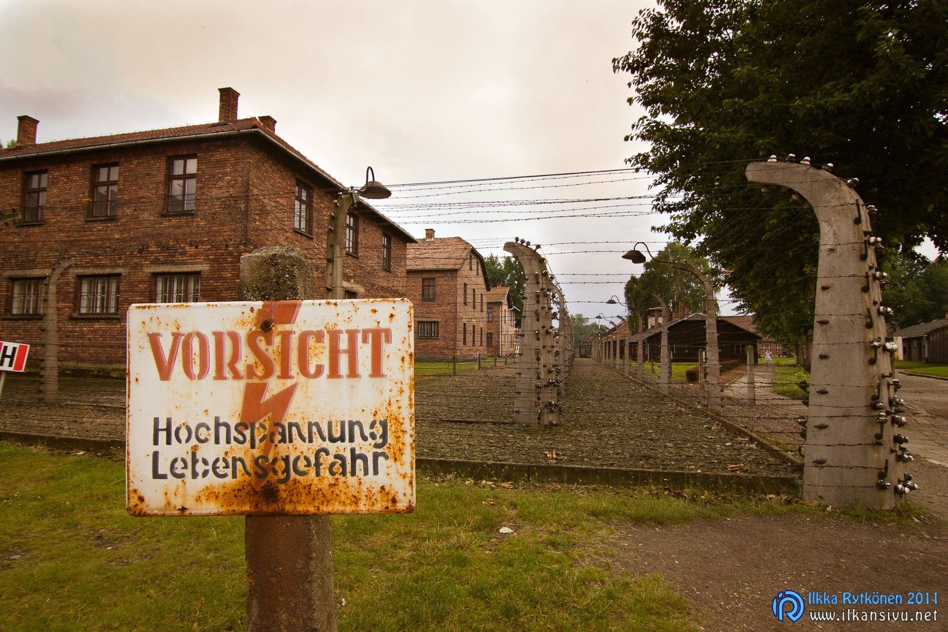 Korkeajännitteestä varoittava kyltti parakkien ja piikkilanka-aidan vieressä Auschwitz I:ssä