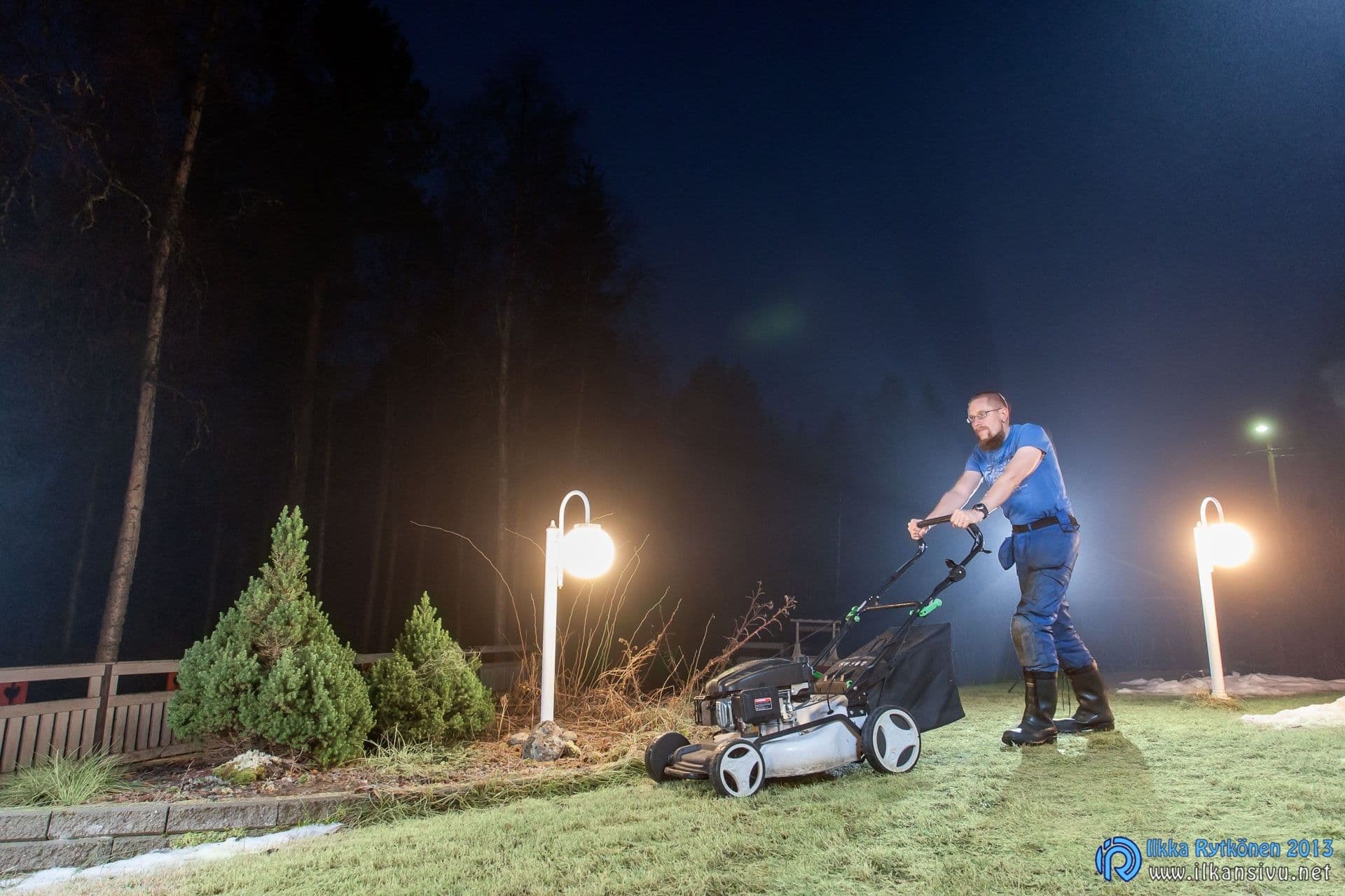 Mies leikkaa nurmikkoa pihavalojen loisteessa yöllä