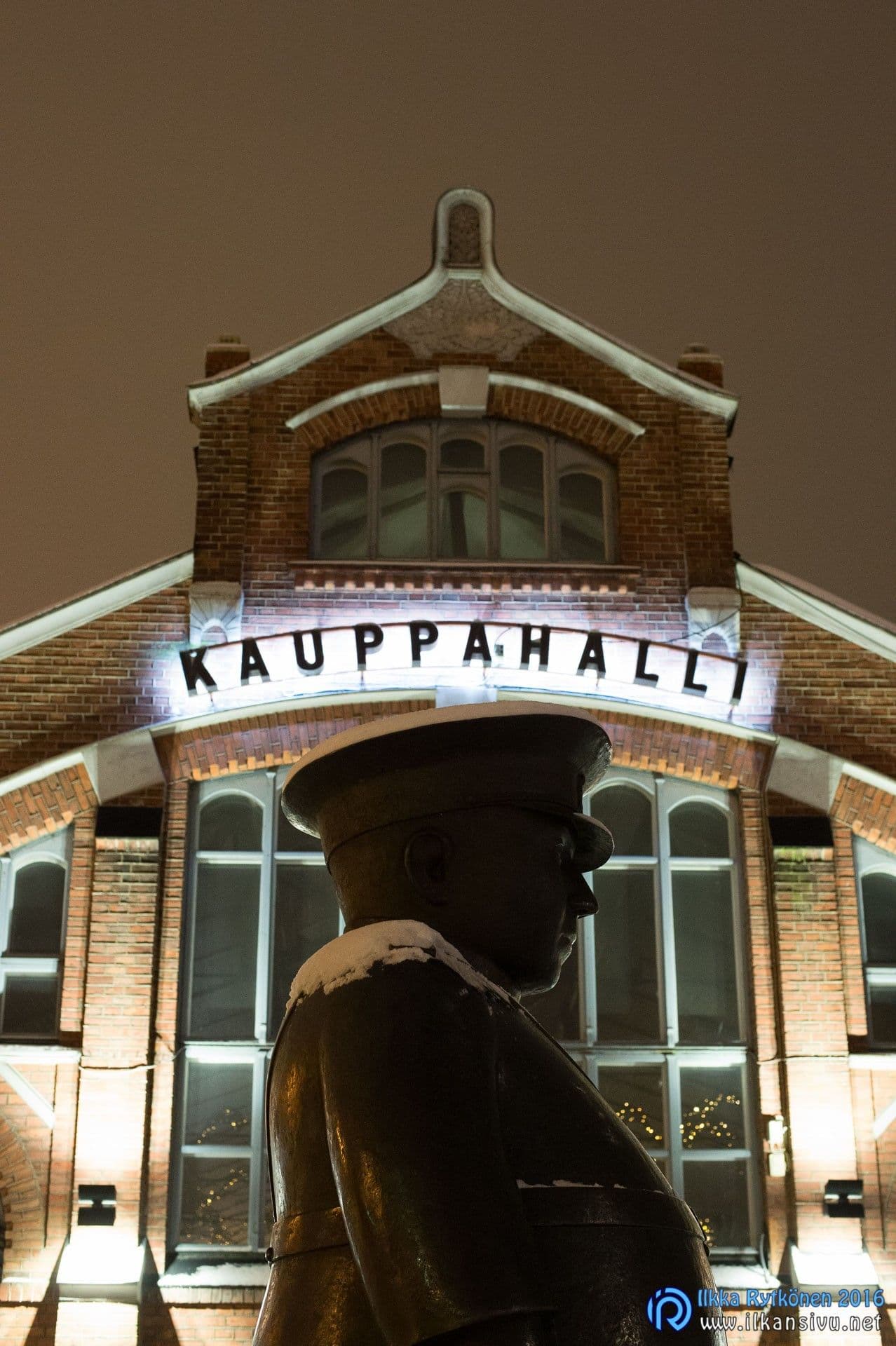 Patsaan tumma siluetti valaistun kauppahallin edessä yökuvassa