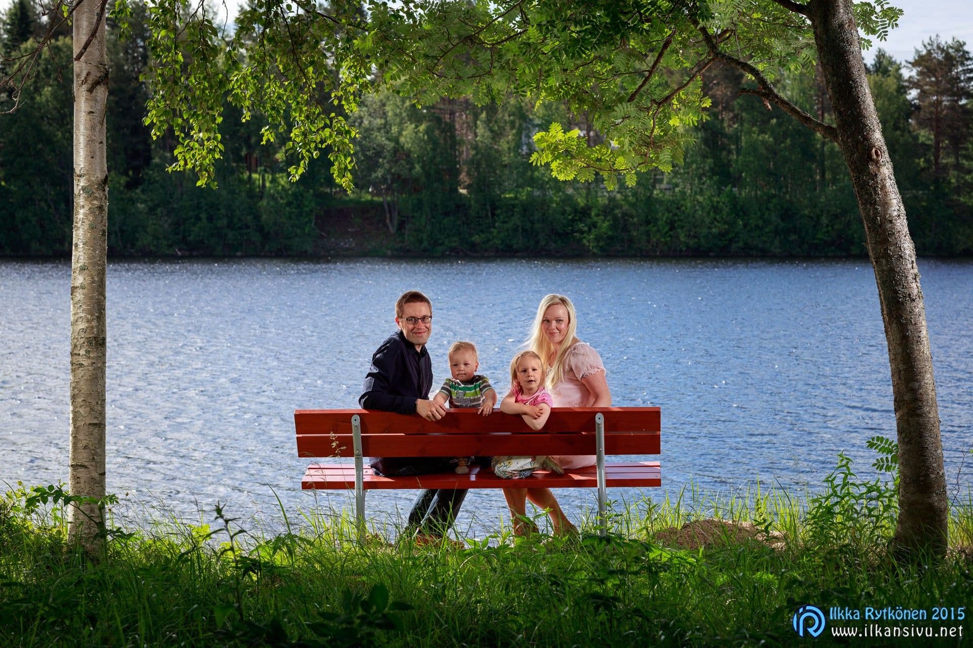 A family sits on a red bench beside the water between trees
