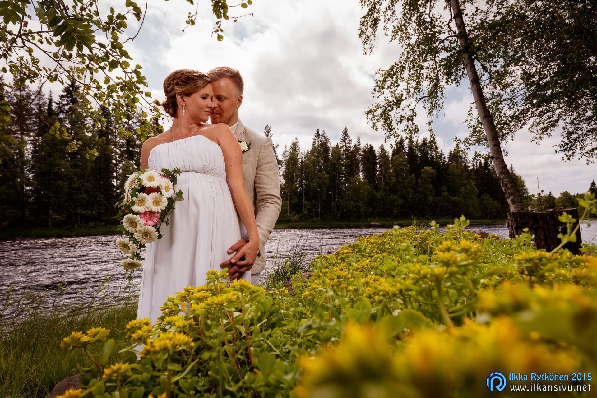 Laura ja Jukka seisovat lähekkäin Kiiminkijoen rannalla keltaisten kukkien keskellä