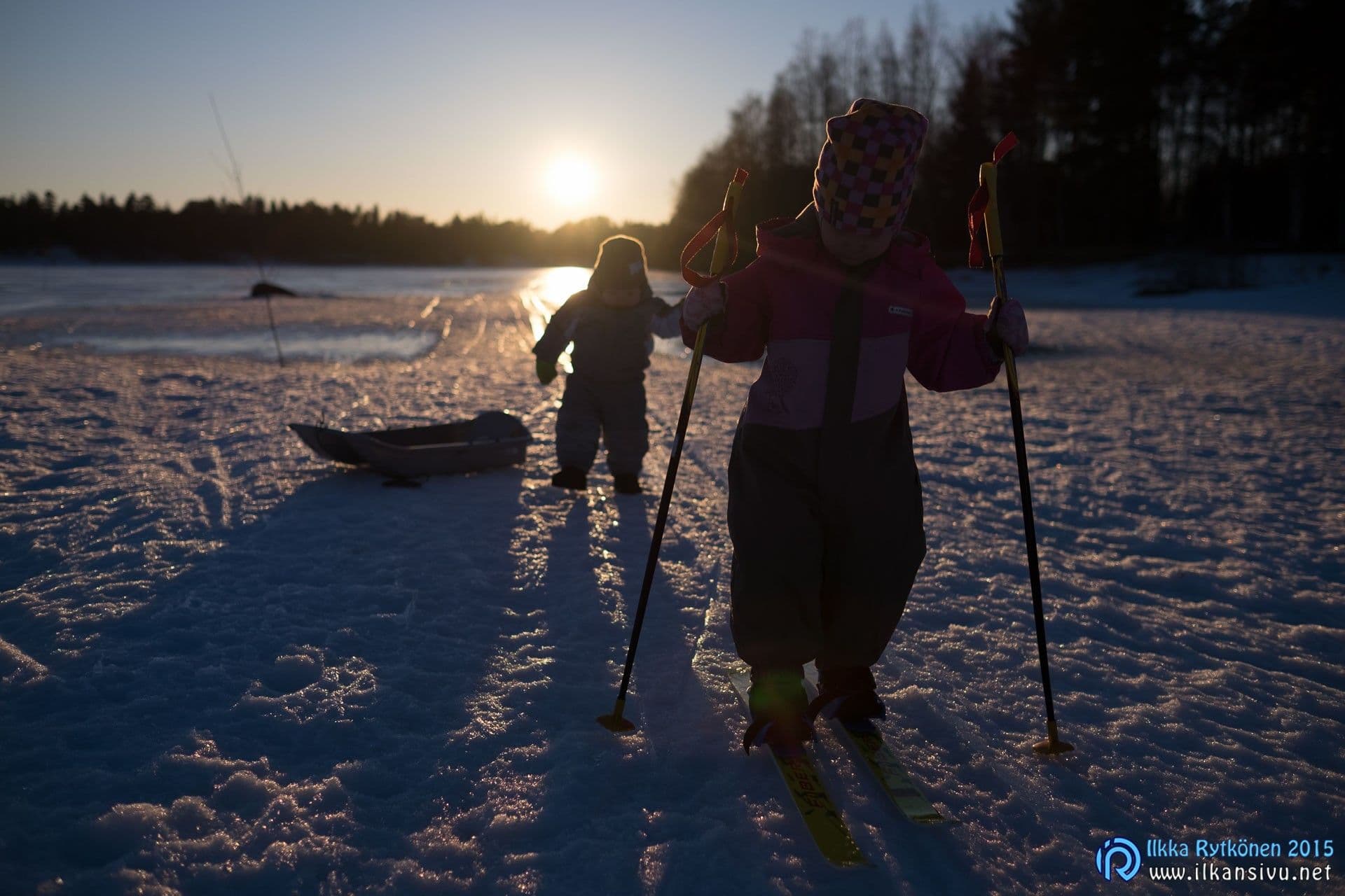 Lapset lumessa matalan auringon vastavalossa, kuvan etuala jää tummaksi